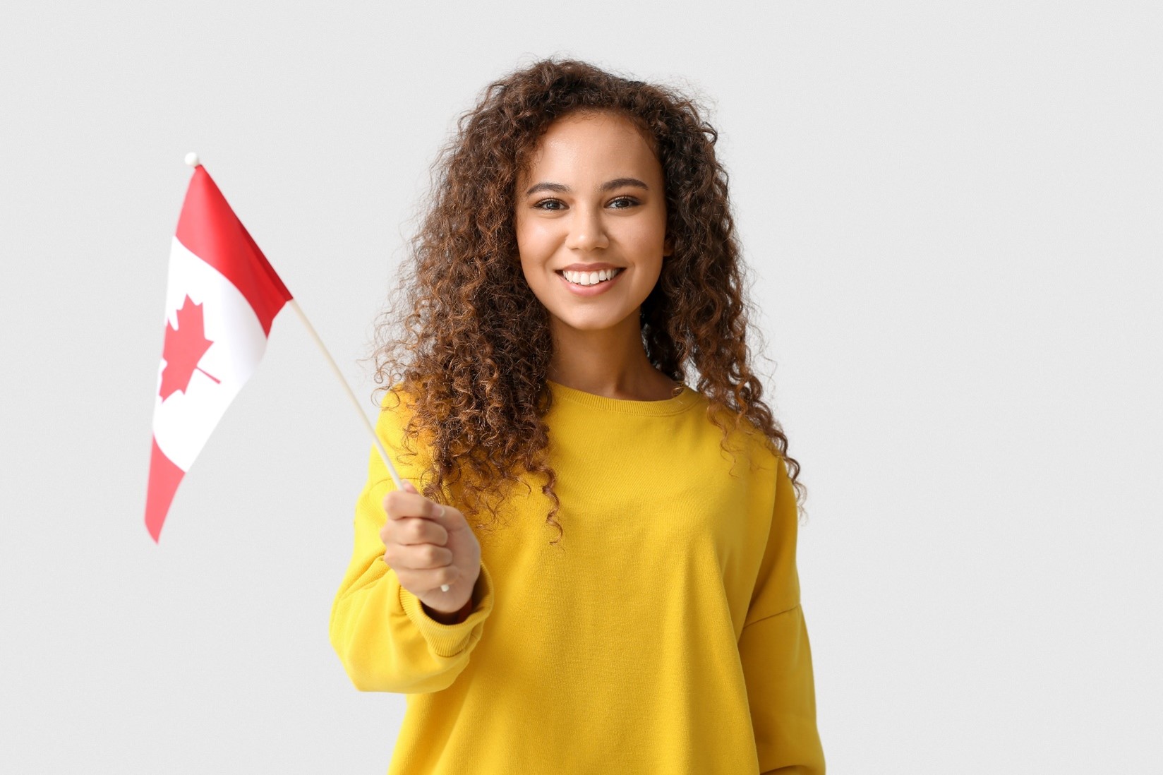 Young woman wearing a yellow sweatshirt and smiling while holding a Candian flag. 